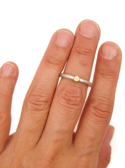 Feiner Ring aus mattiertem Silber mit einer zarten Goldfassung, die einen kleinen Brillanten hält. Die klaren Proportionen und das Zusammenspiel aus kühlem Silber und warmem Gold stehen im Fokus der Darstellung.